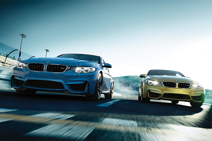 Mood shot of a blue BMW M3 and yellow BMW M4 side by side on a race track. 